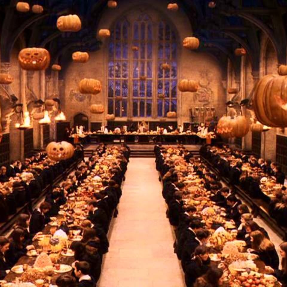 A grand hall with long tables filled with people, decorated with floating jack-o'-lanterns.