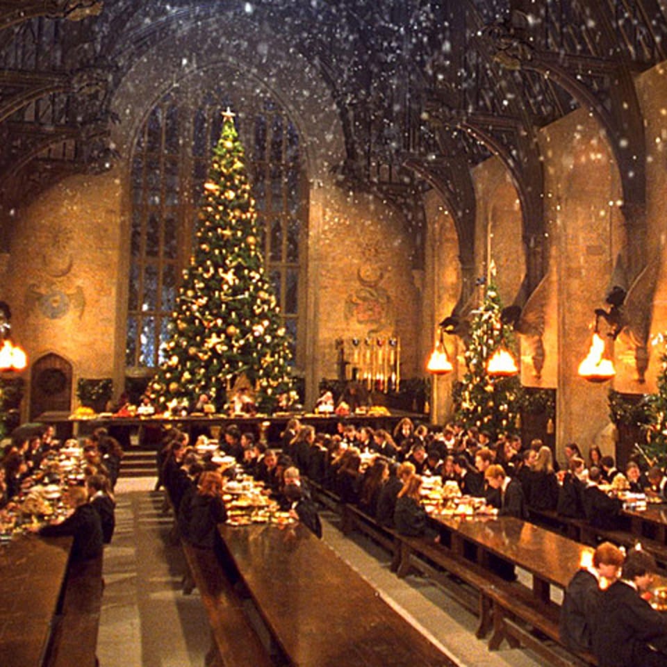 Students in robes dine at long tables in a grand hall decorated with Christmas trees and falling snow.